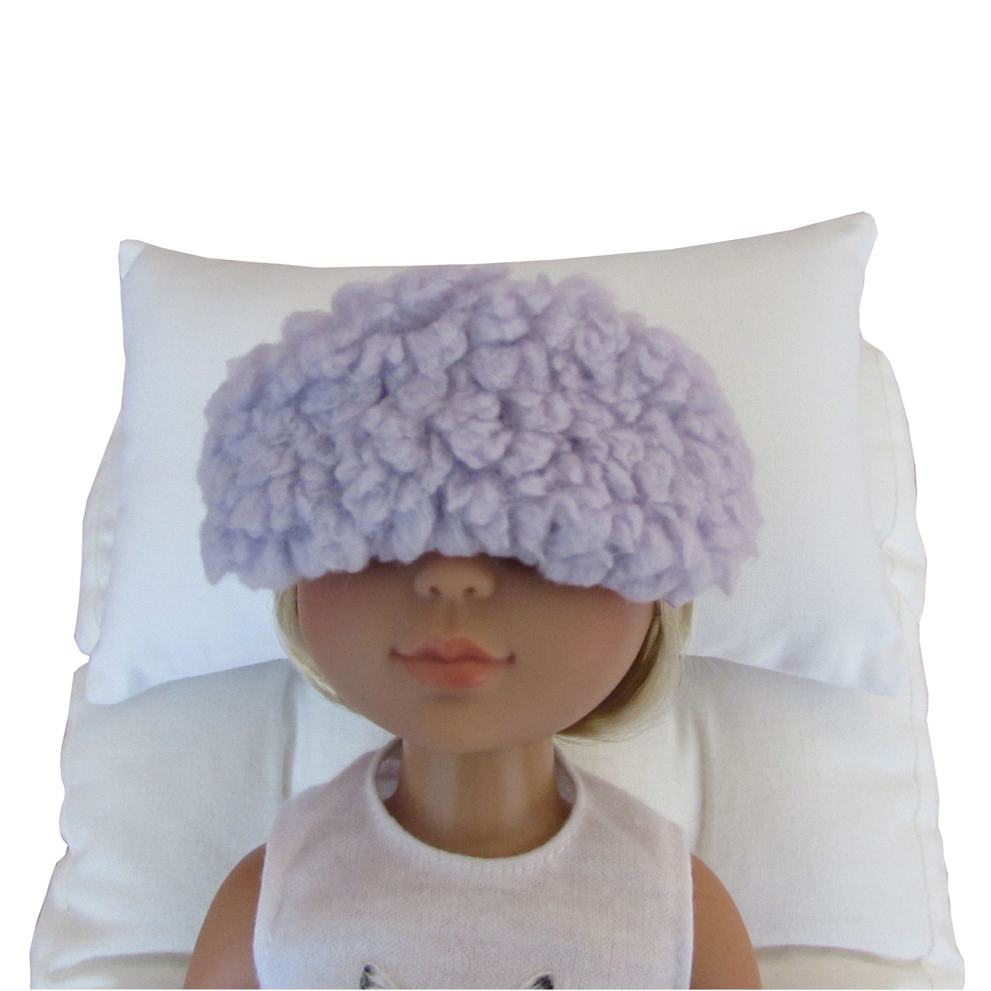 A doll with blonde hair wearing a lilac-colored fuzzy sleep mask, resting on a white pillow against a white background.
