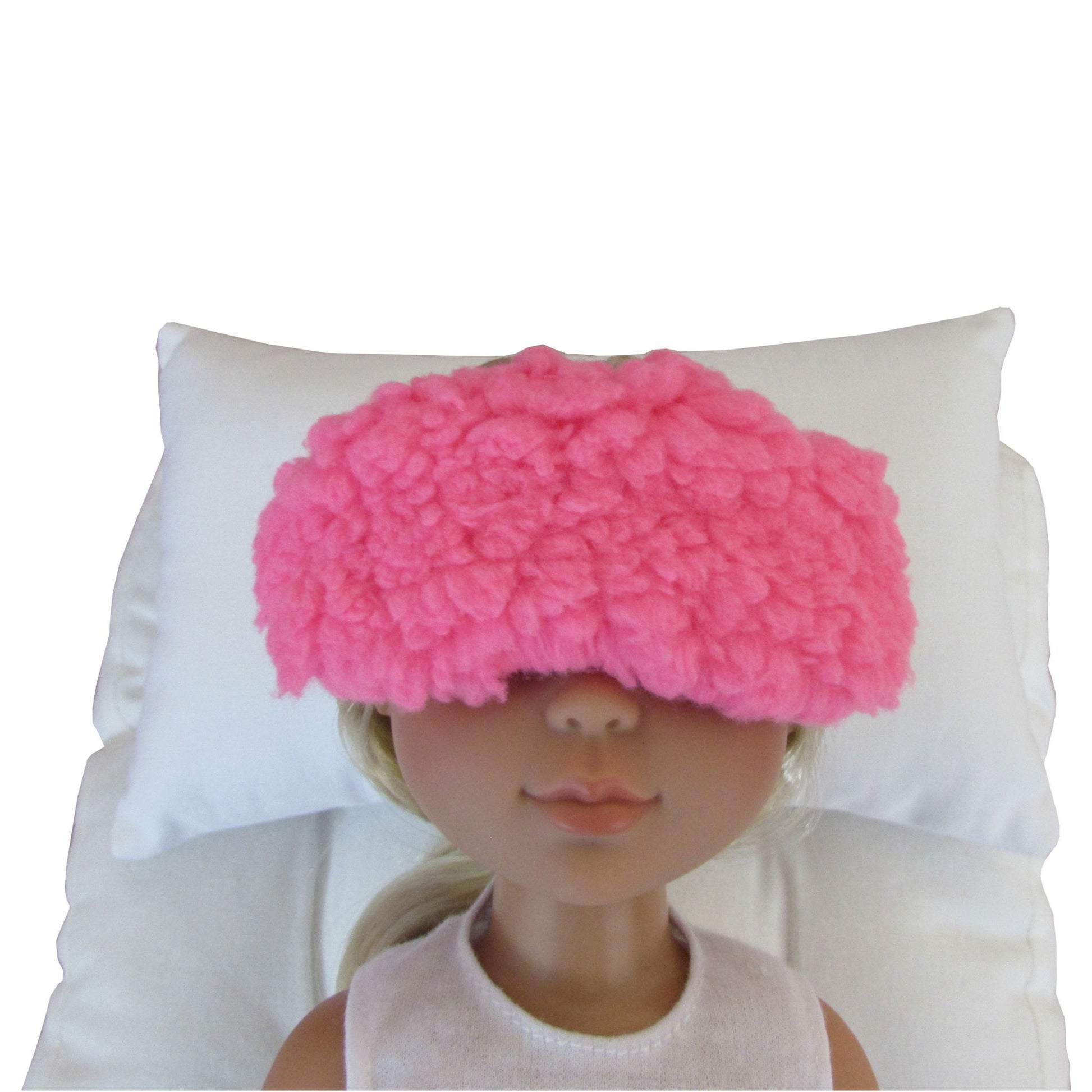 A pink fuzzy sleep mask worn by a doll, resting on a white pillow against a white background.