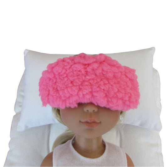 A pink fuzzy sleep mask worn by a doll, resting on a white pillow against a white background.