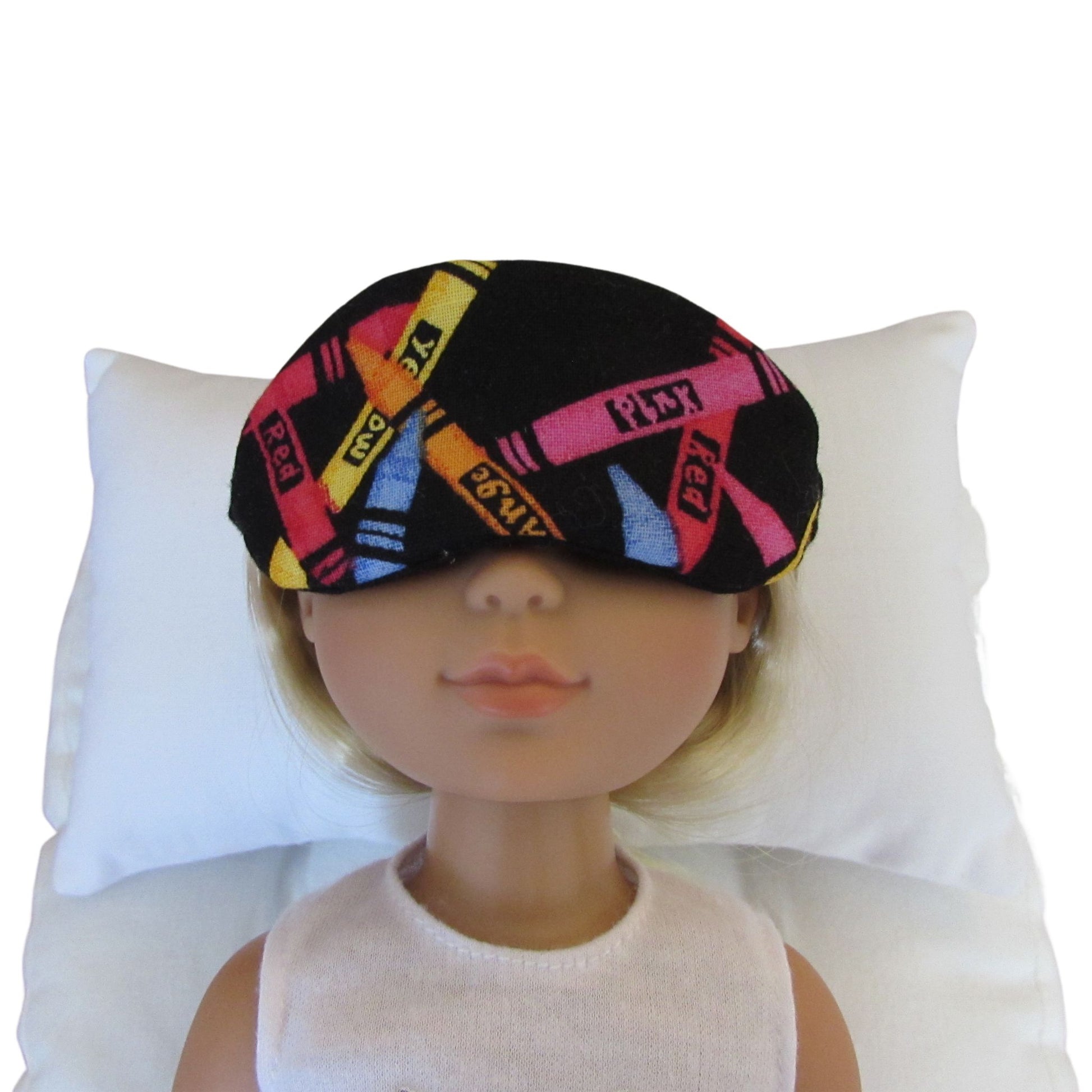 A doll with blonde hair wearing a black sleep mask patterned with colorful crayons, resting on a white pillow against a white background.