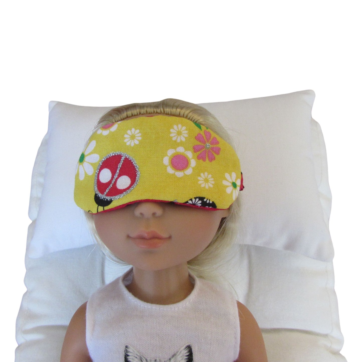 A doll wearing a yellow sleep mask with a ladybug and daisy pattern, resting on a white pillow.