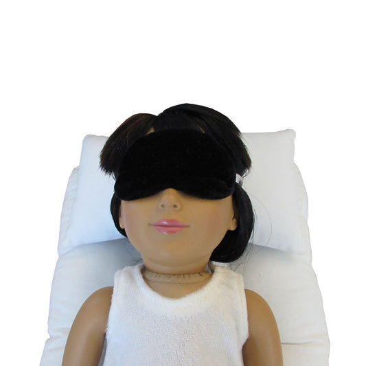 An 18-inch doll is wearing a black velvet sleep mask while lying on a white doll pillow and a white doll mattress.