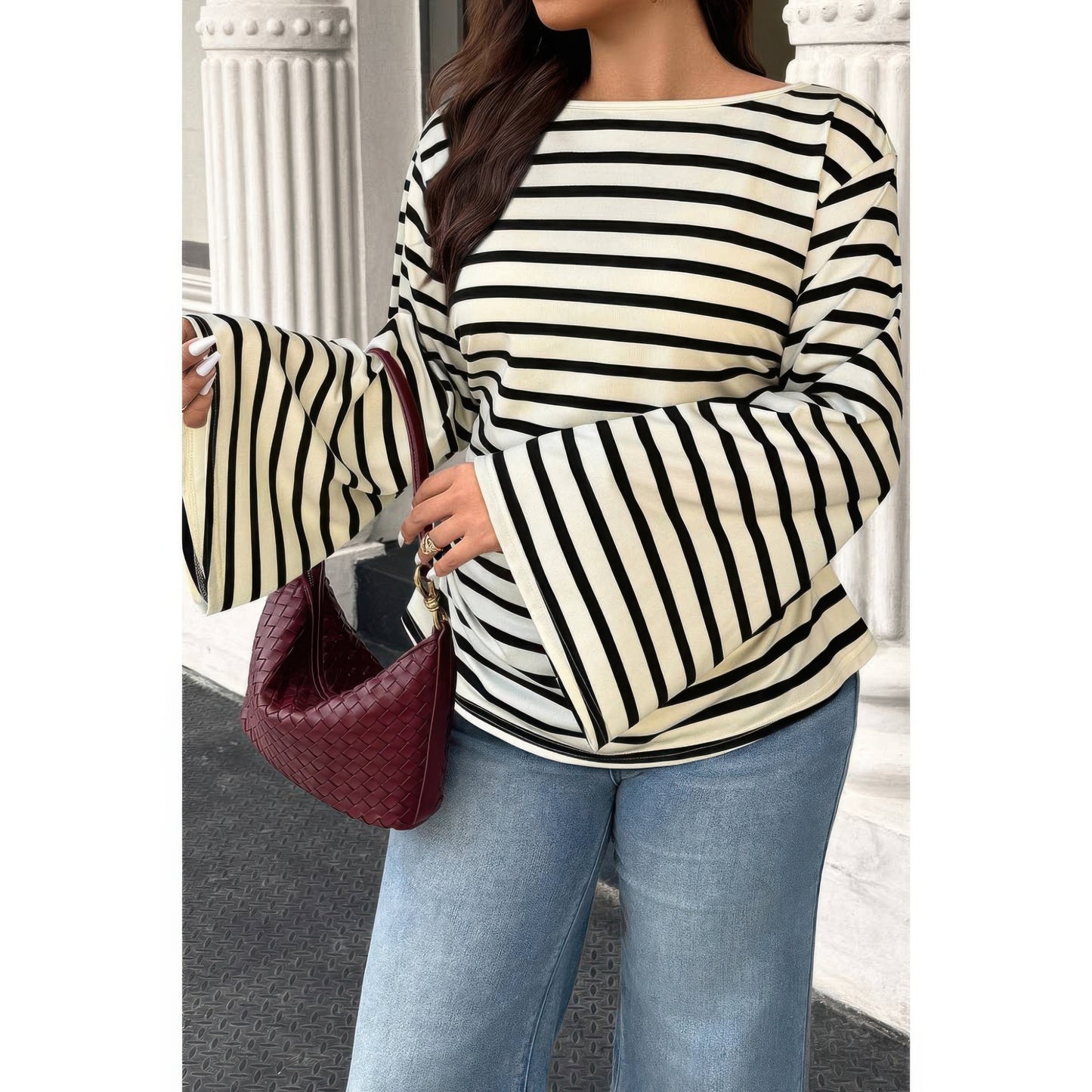 A woman wearing a cream and black horizontally striped long-sleeve knit top with flared bell sleeves, paired with light blue jeans and carrying a burgundy woven handbag.