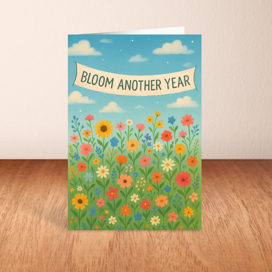 Greeting card with colorful flowers and 'Bloom Another Year' text on a wooden surface.