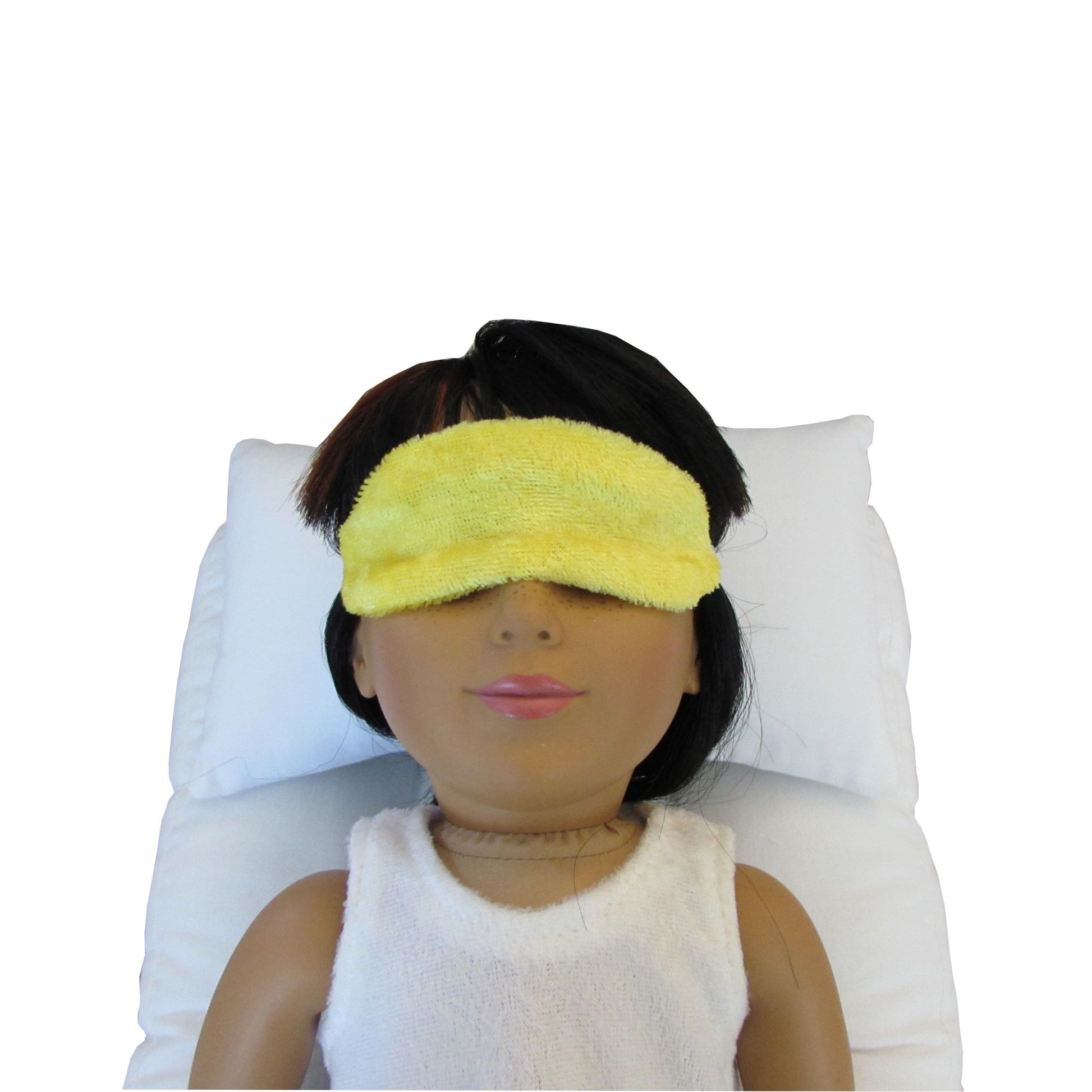 An 18-inch doll is wearing a bright yellow panne velvet sleep mask while lying on a white doll pillow and doll mattress.