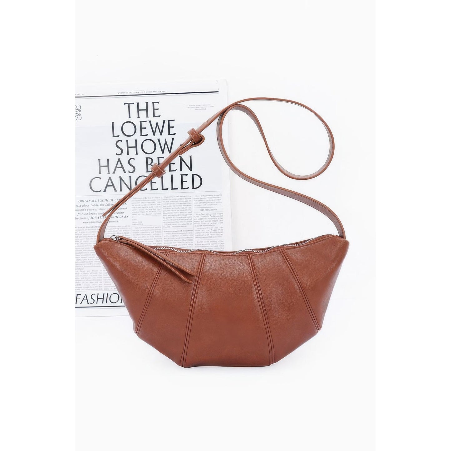 A brown faux-leather crescent shoulder bag with a curved strap, displayed on a white background with a printed magazine page behind it.