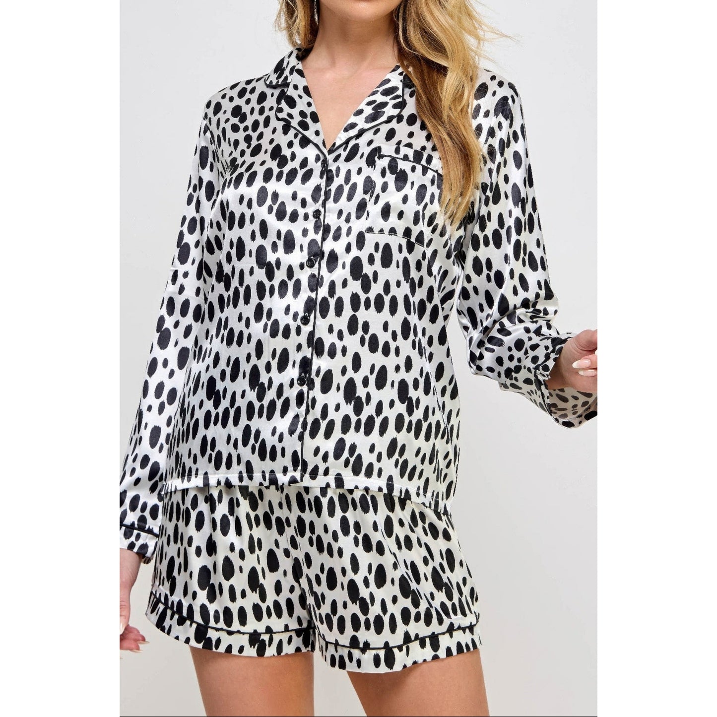 A woman wearing a matching long-sleeve button-front pajama top and shorts set in a black and white spotted animal print, photographed against a light gray studio background.