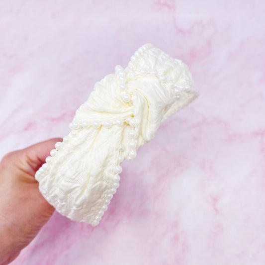 White knitted headband held by a hand against a pink background