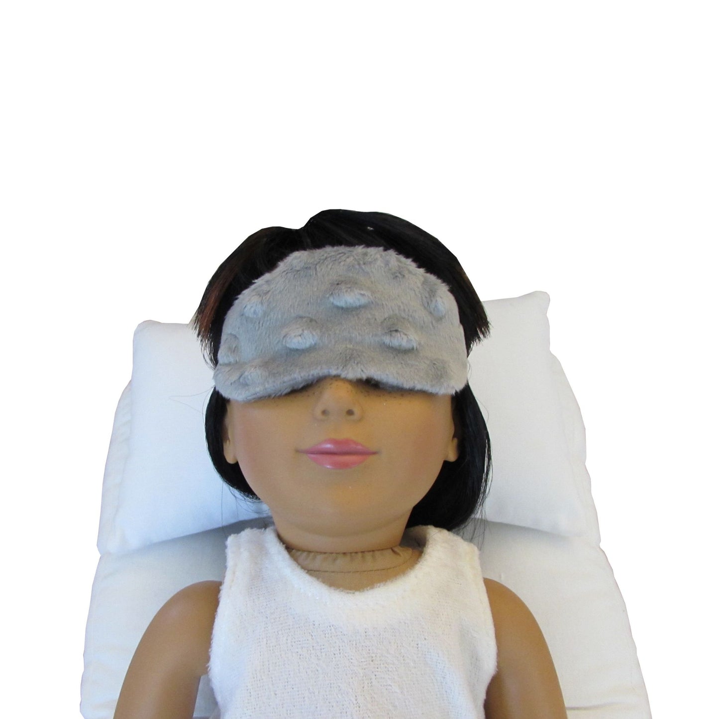 An 18-inch doll is wearing a gray minky sleep mask while lying on a white doll pillow and a white doll mattress.