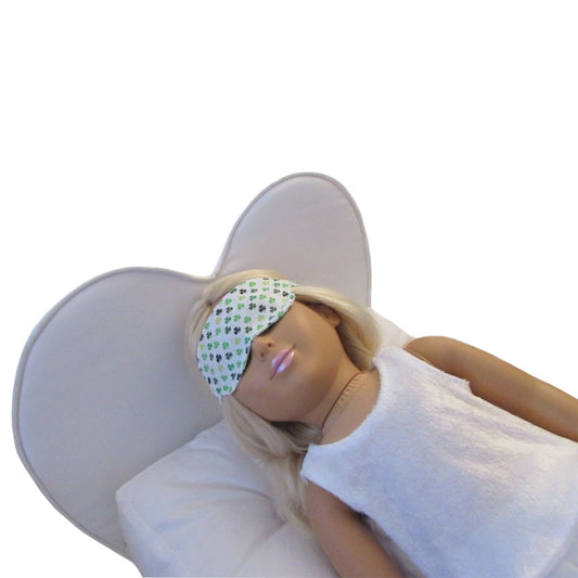 An 18-inch doll wearing a green clover print sleep mask while lying on a white heart doll bed.
