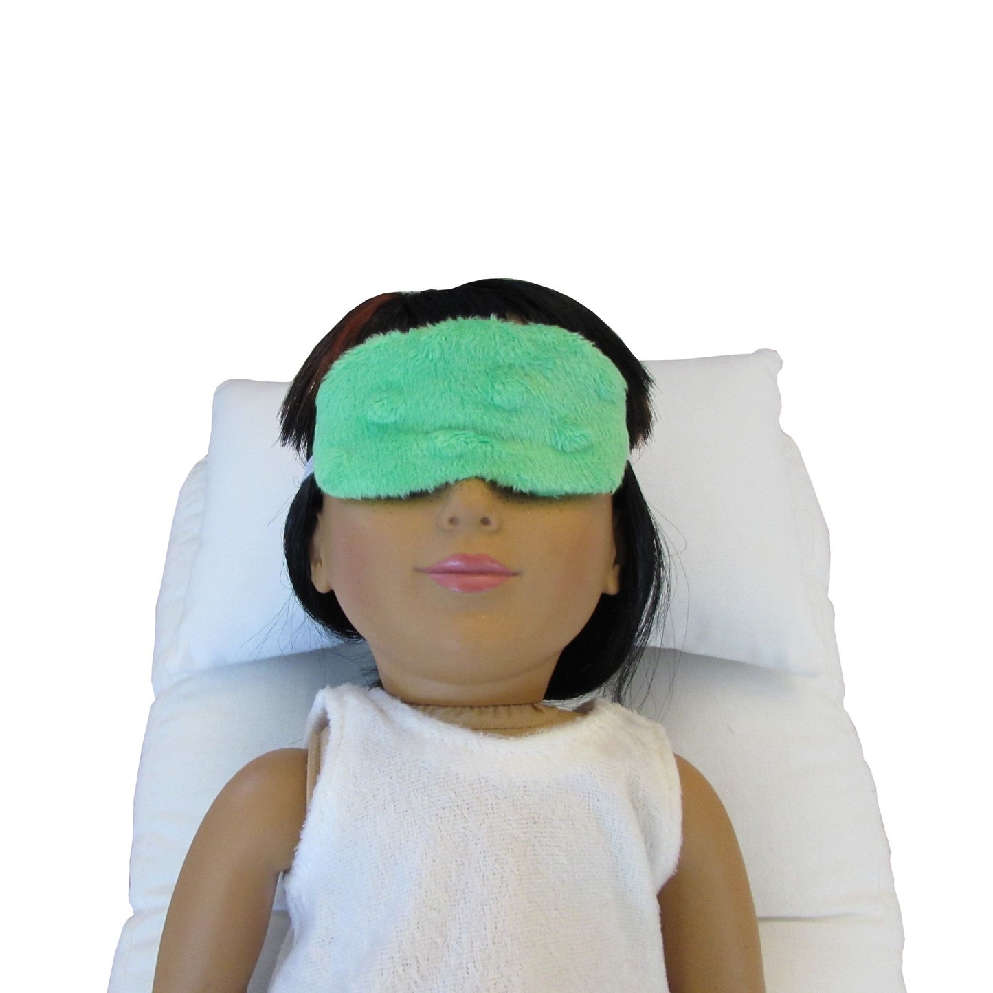 An 18-inch doll is wearing a green minky sleep mask while lying on a white doll pillow and white doll mattress.