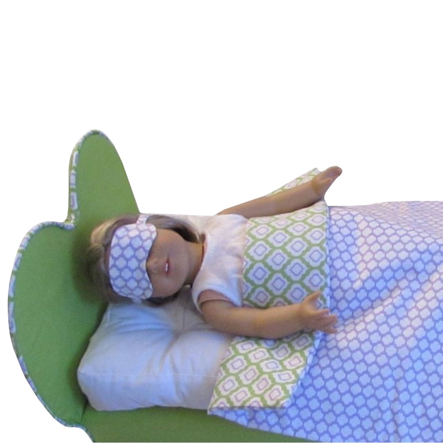 Doll lying on a green pillow with a patterned blanket and sleep mask on a white background