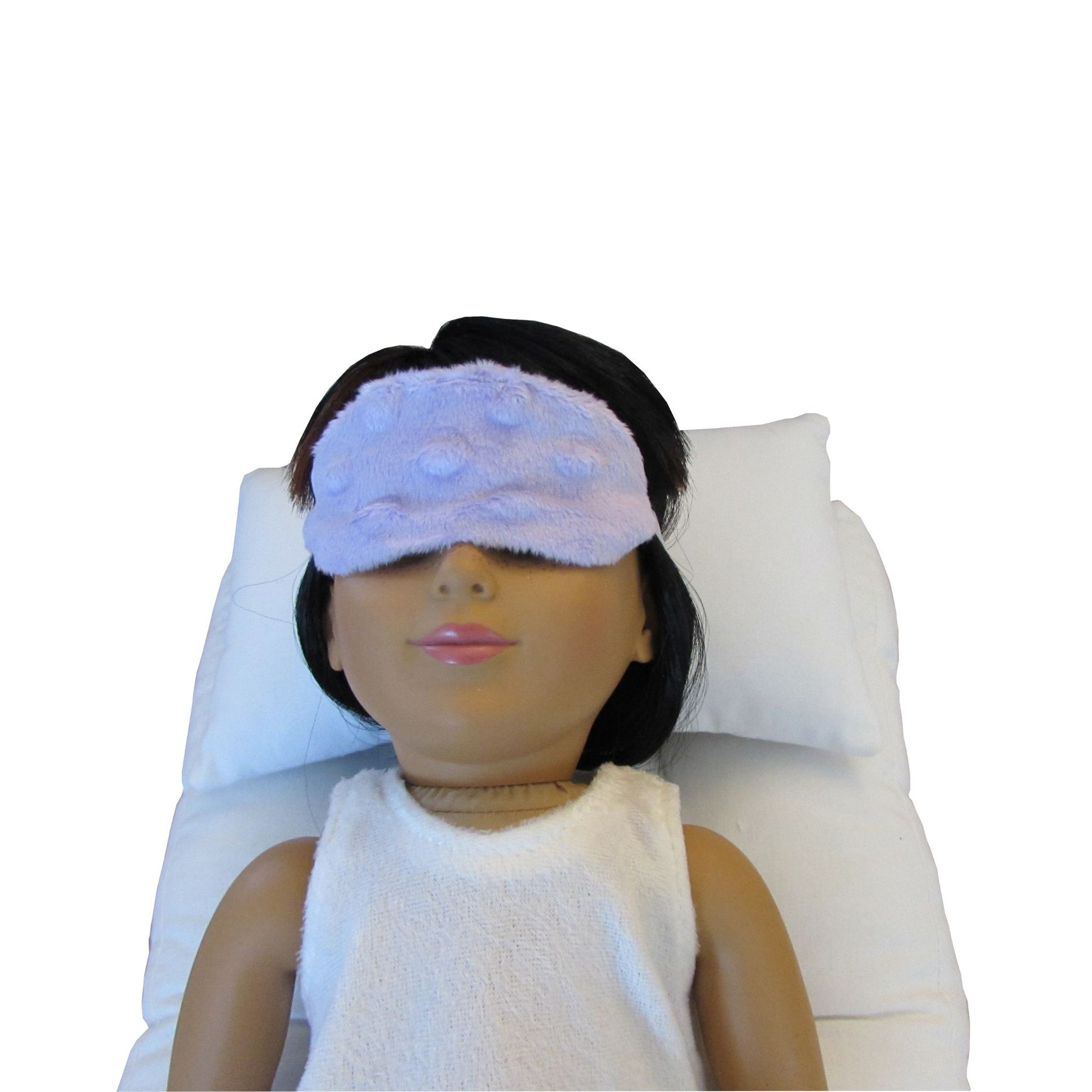 An 18-inch doll is wearing a lavender minky sleep mask while lying on a white doll pillow and a white doll mattress.