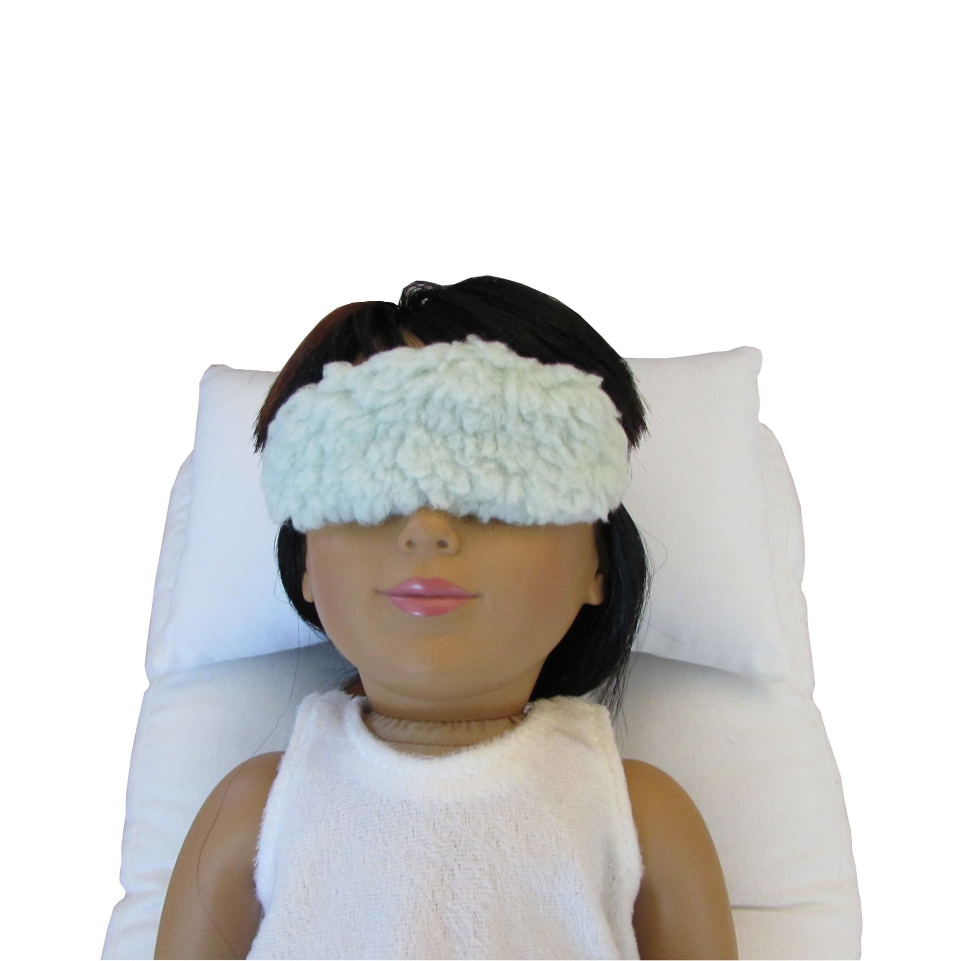 An 18-inch doll is wearing a light green faux sherpa fur doll sleep mask while lying on a white doll pillow and a white doll mattress.
