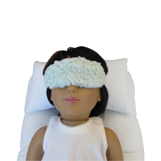 An 18-inch doll is wearing a light green faux sherpa fur doll sleep mask while lying on a white doll pillow and a white doll mattress.