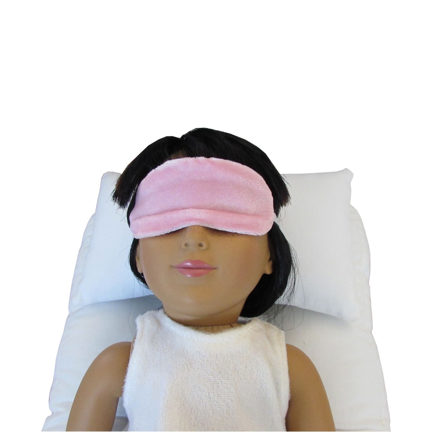 An 18-inch doll is wearing a light pink stretch velvet sleep mask while lying on a white doll  pillow and a white doll mattress.