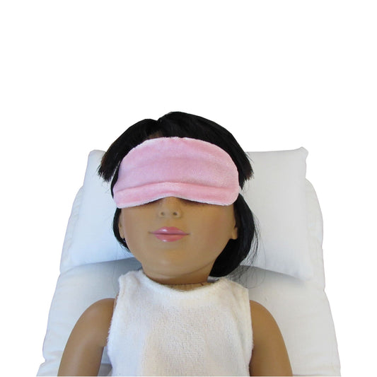 An 18-inch doll is wearing a light pink stretch velvet sleep mask while lying on a white doll  pillow and a white doll mattress.