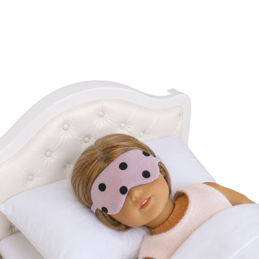 An 18-inch doll wearing a light pink sleep mask with brown dots while lying on a white doll bed.