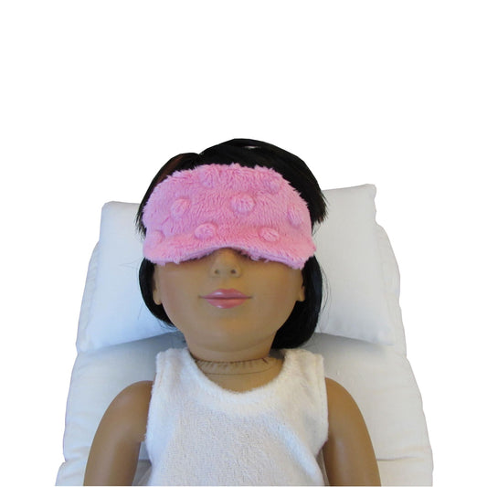 An 18-inch doll is wearing a light pink minky sleep mask while lying on a doll pillow and doll mattress.