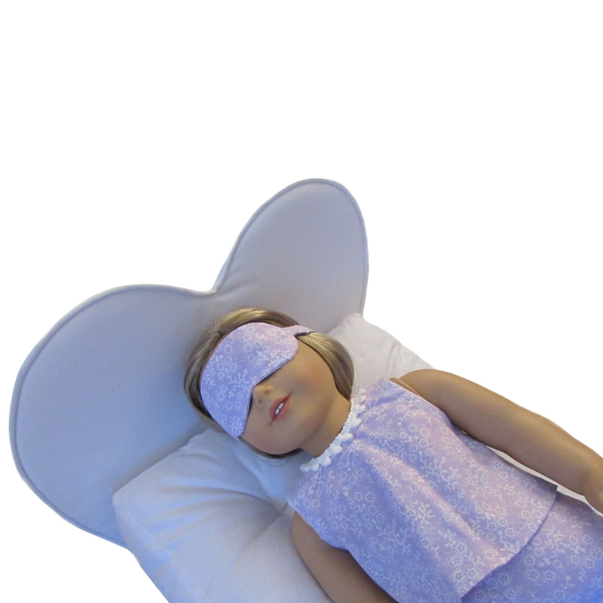 An 18-inch doll wearing a lilac and white floral sleep mask while lying on a white heart doll bed.