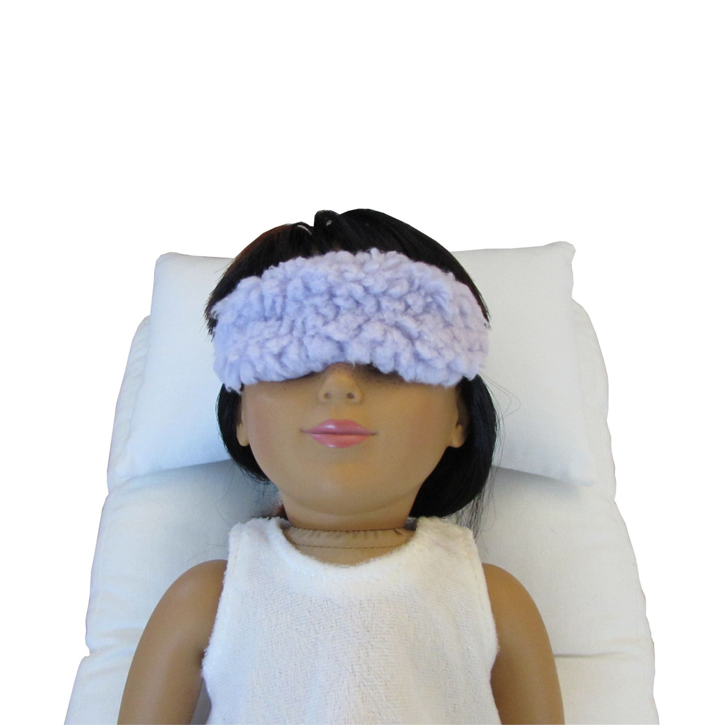 An 18-inch doll is wearing a lilac faux sherpa fur doll sleep mask while lying on a white doll pillow and a white doll mattress.
