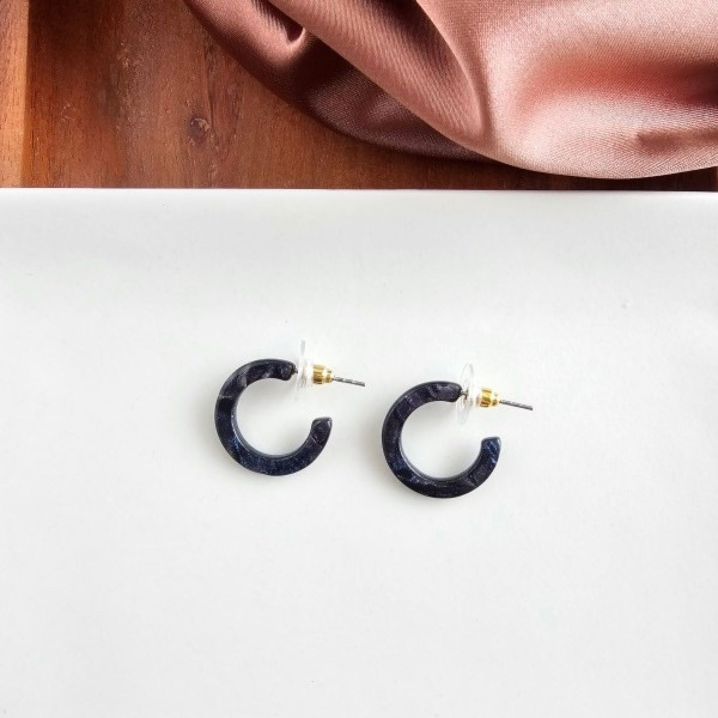 Pair of dark blue hoop earrings on a white surface with a pink fabric background