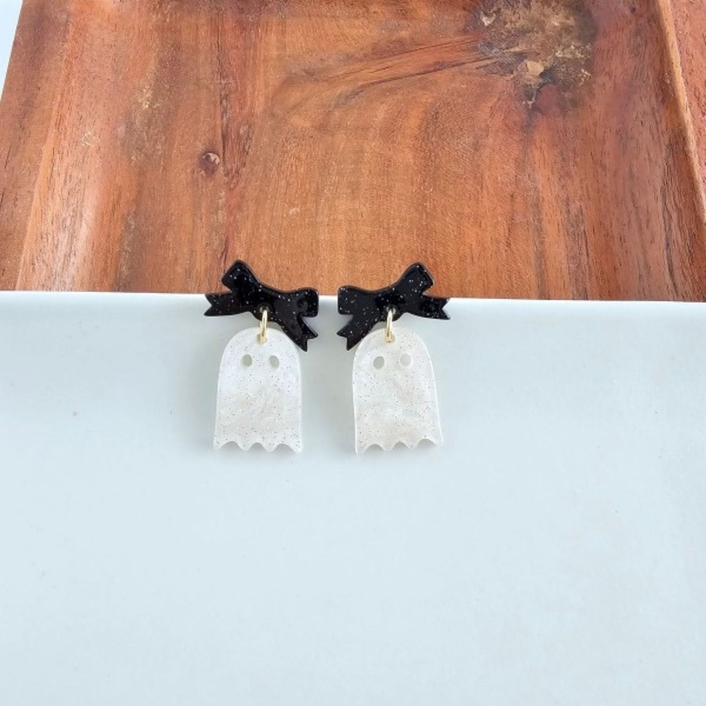 Ghost-shaped earrings with black bows on a white surface and wooden background