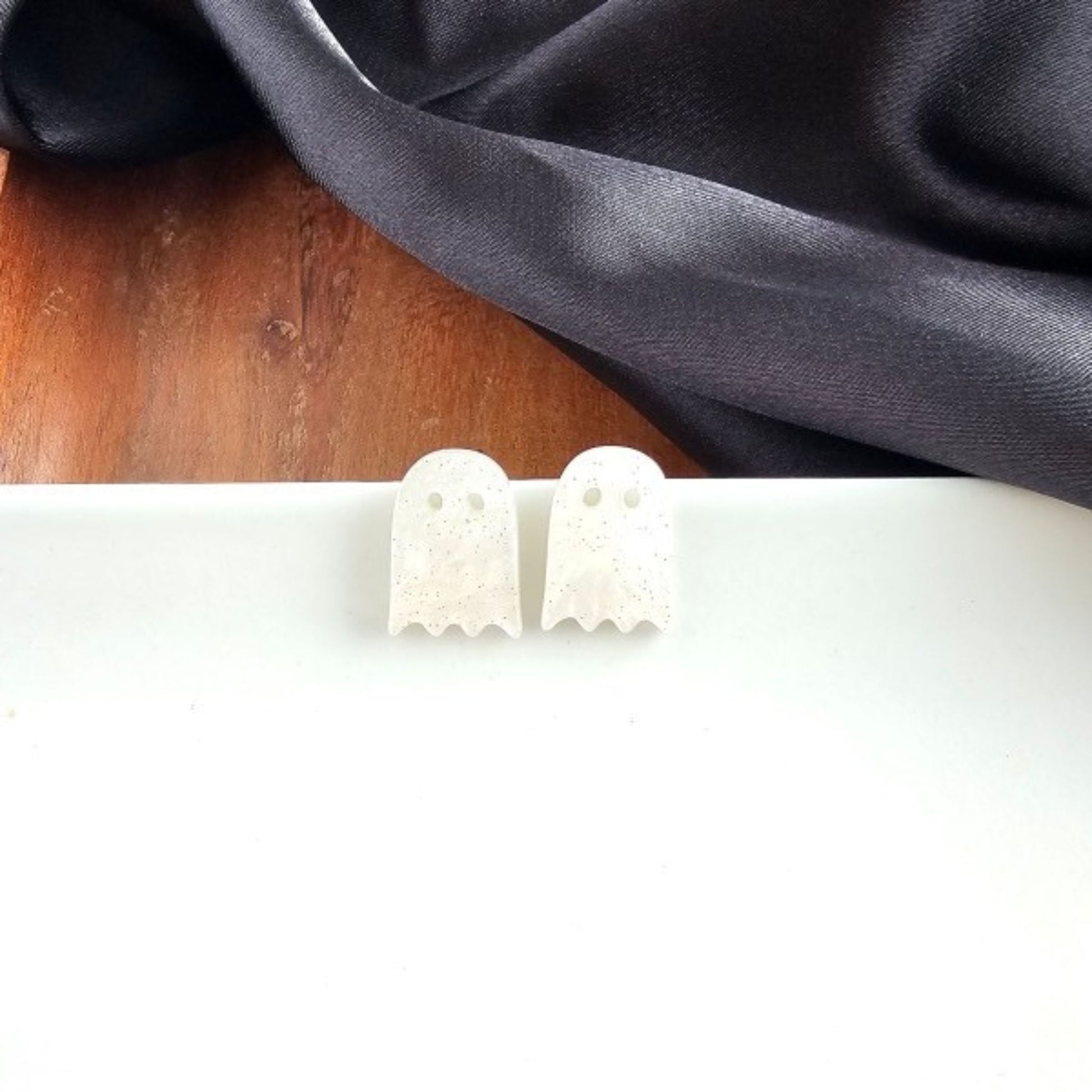 White ghost-shaped earrings on a white surface with a dark fabric background