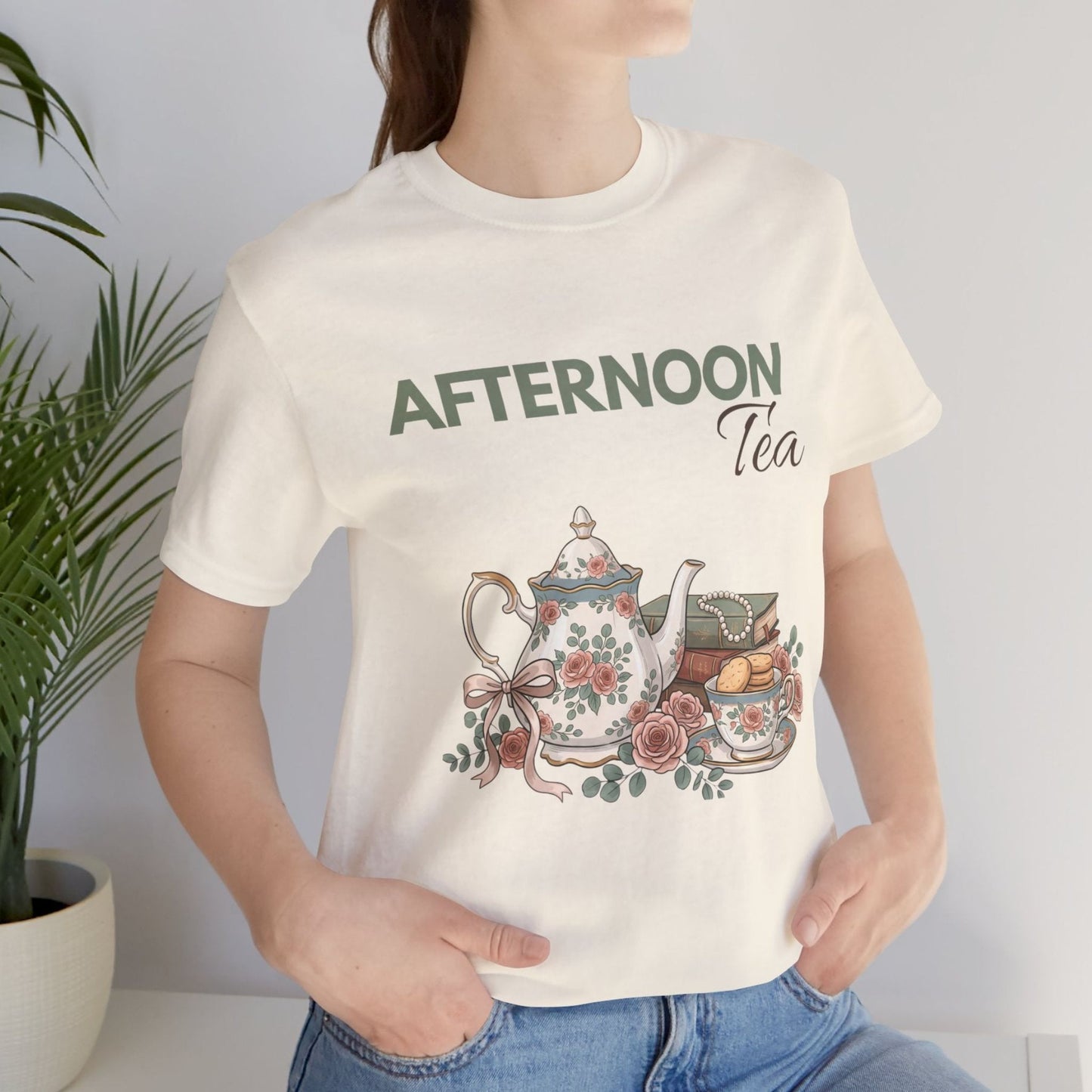 A woman standing by a plant with a natural colored t-shirt with 'AFTERNOON Tea' on the front.