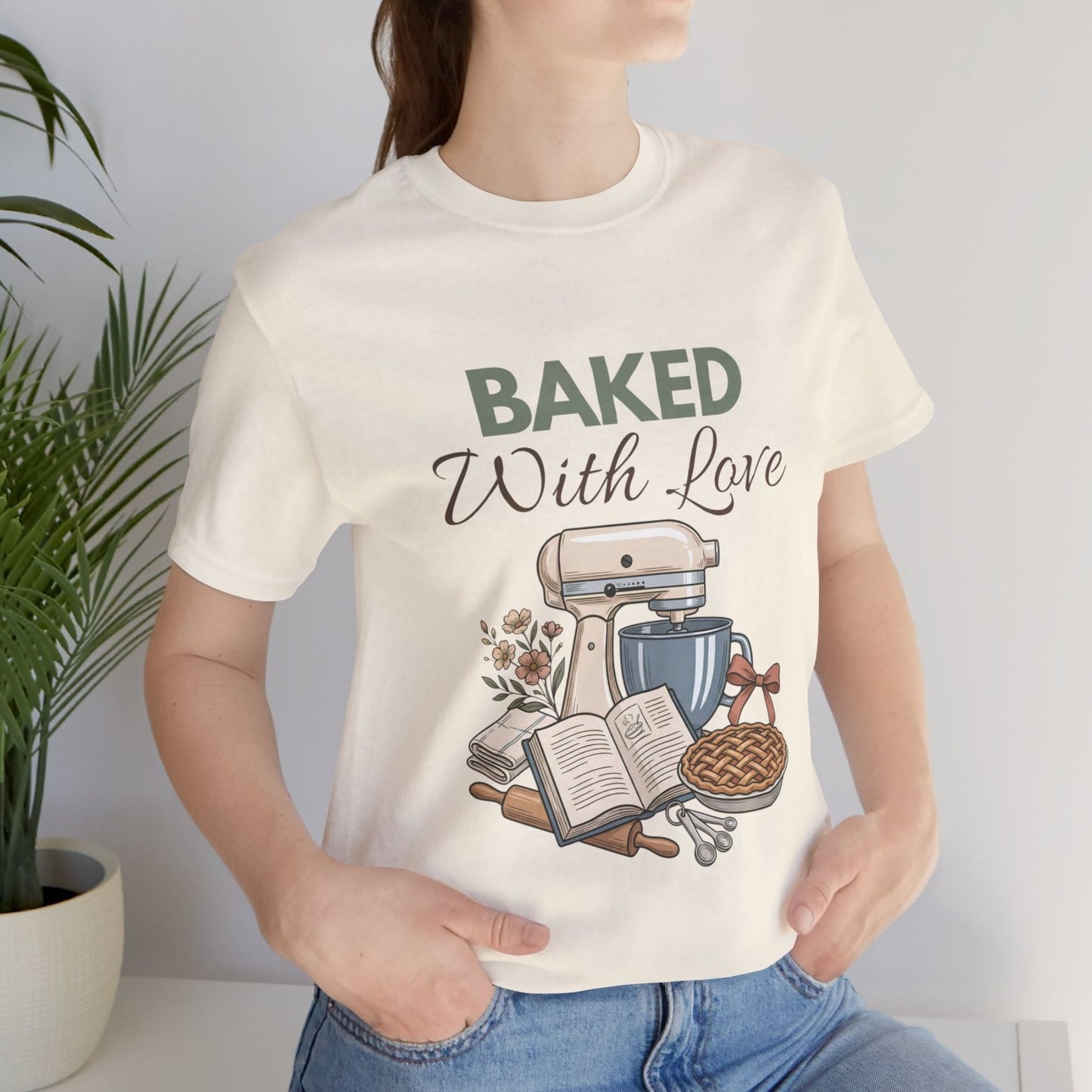 A woman wearing a natural colored t-shirt with 'BAKED With Love' on the front and standing by a plant.