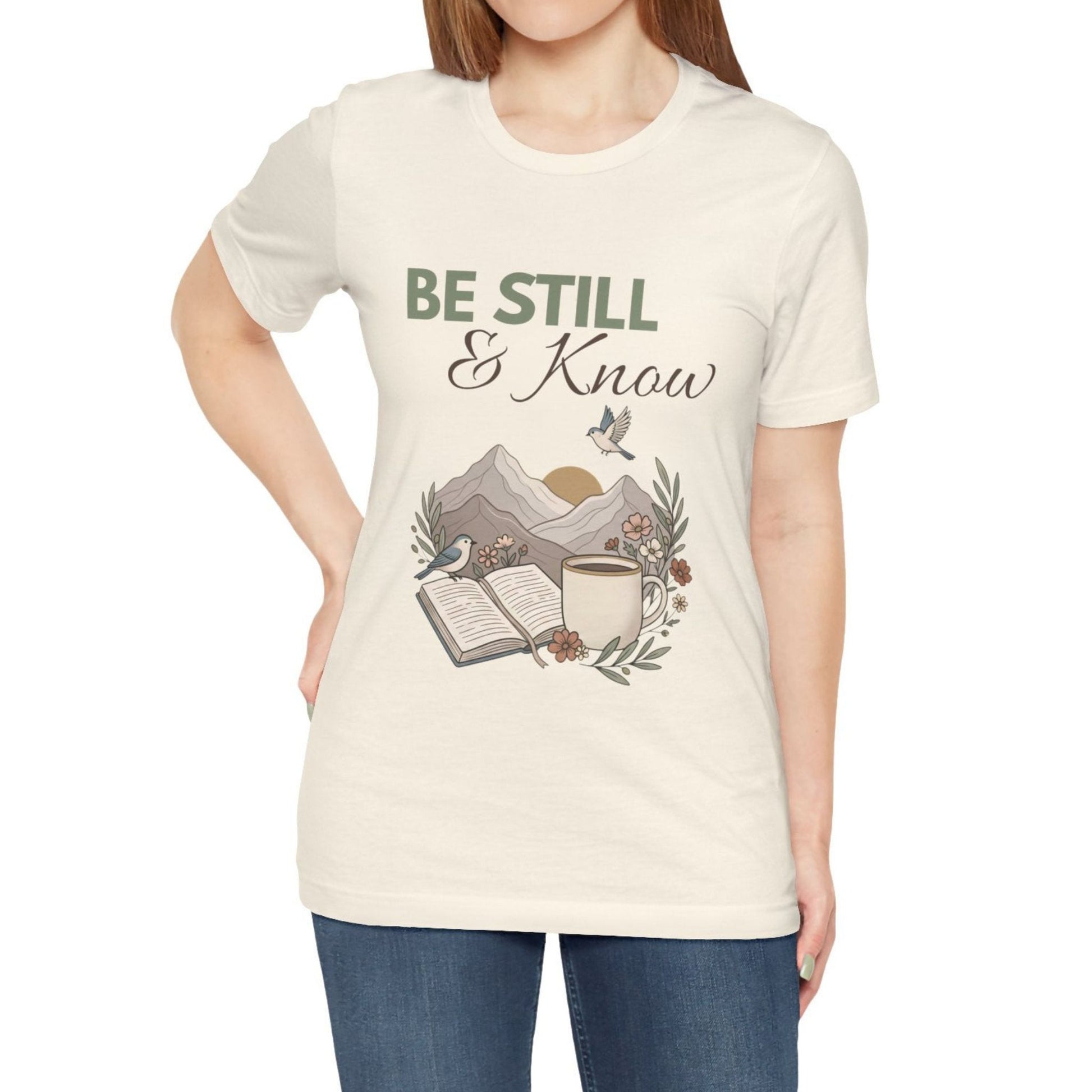Person wearing a natural colored t-shirt with 'Be Still & Know' design on a plain background