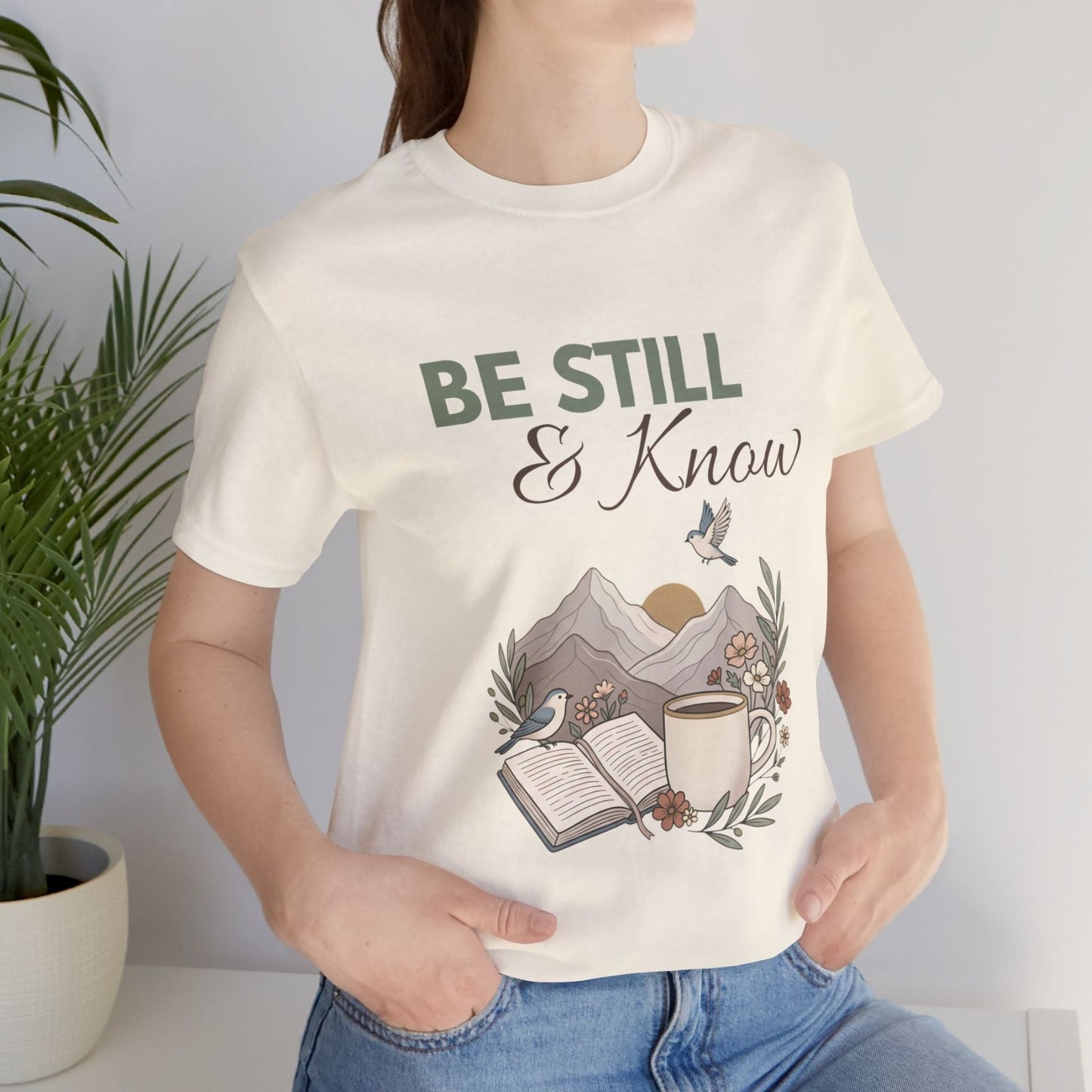Person wearing a natural colored t-shirt with 'Be Still & Know' design, featuring nature elements.