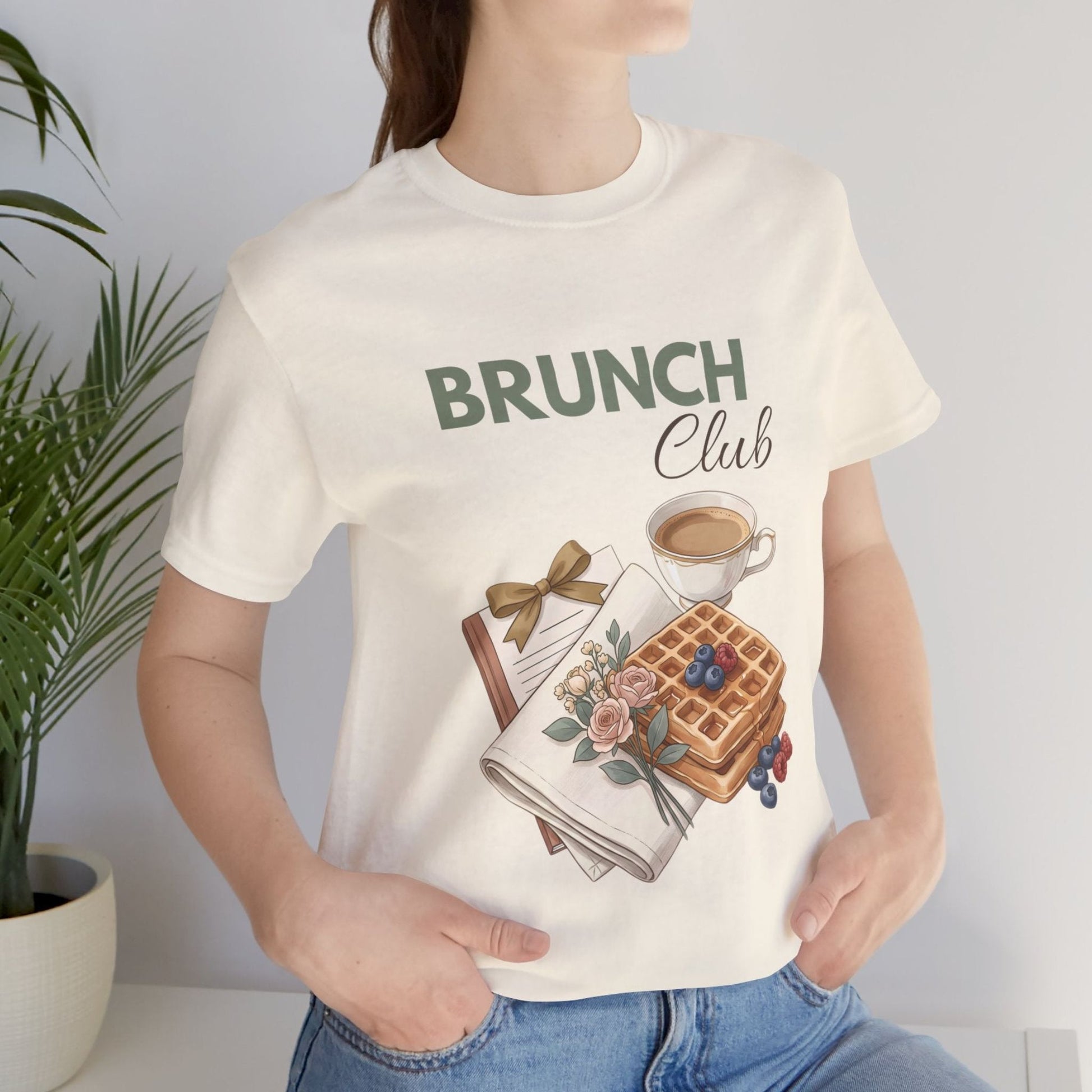 A woman is standing by a plant while wearing a natural colored t-shirt with 'BRUNCH Club' on the front.