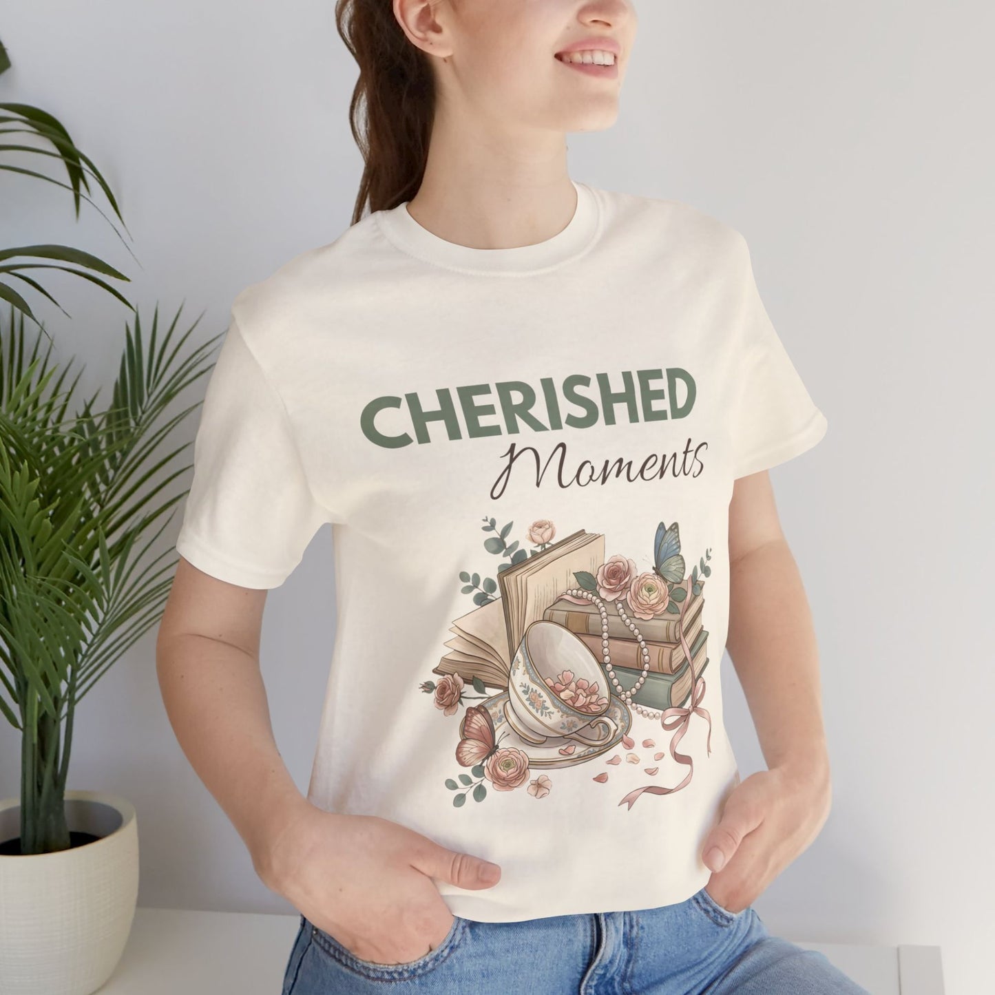 A woman is standing by a plant while wearing a natural colored t-shirt with 'CHERISHED Moments' on the front.