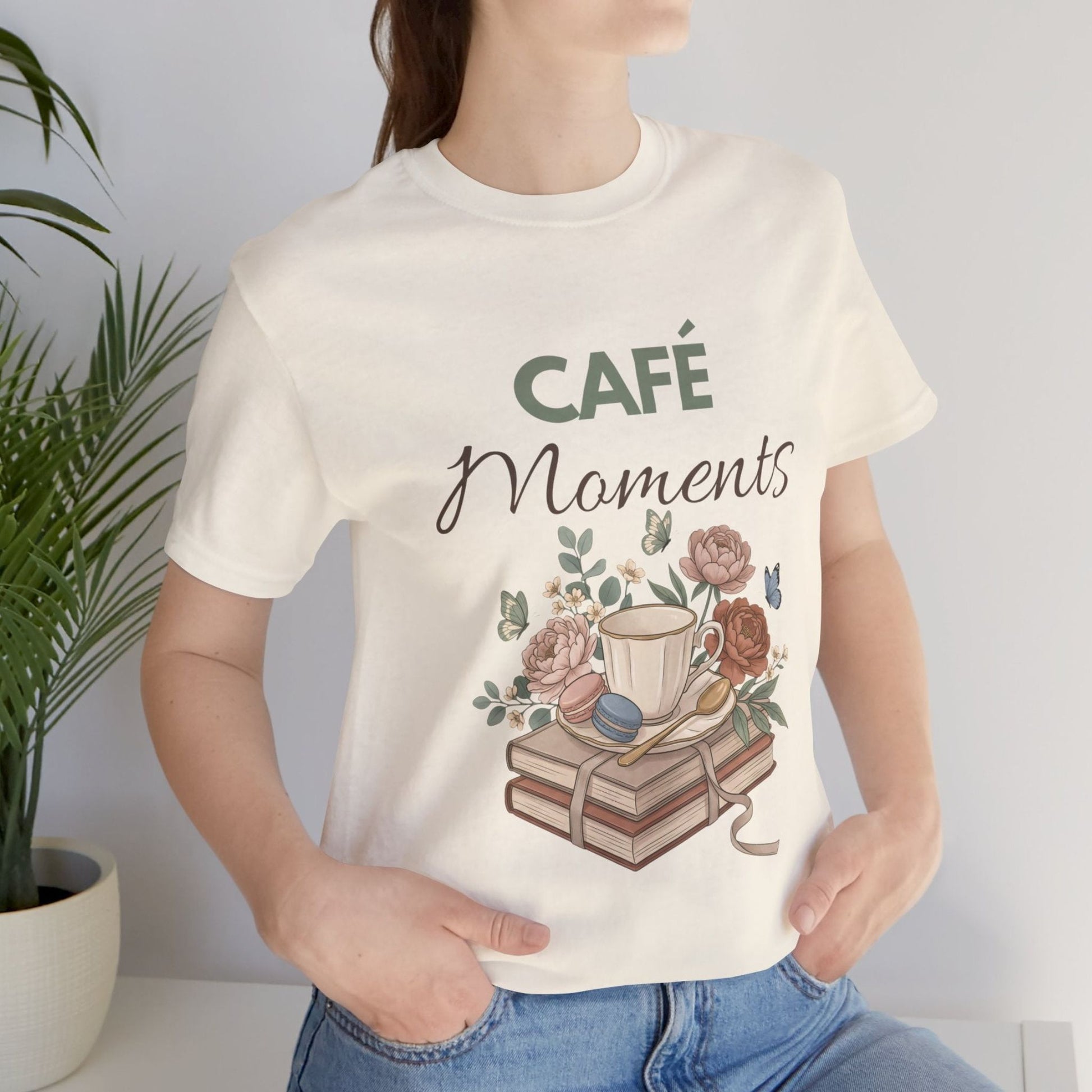 Person wearing a natural colored t-shirt with 'Café Moments' design featuring a coffee cup and books.