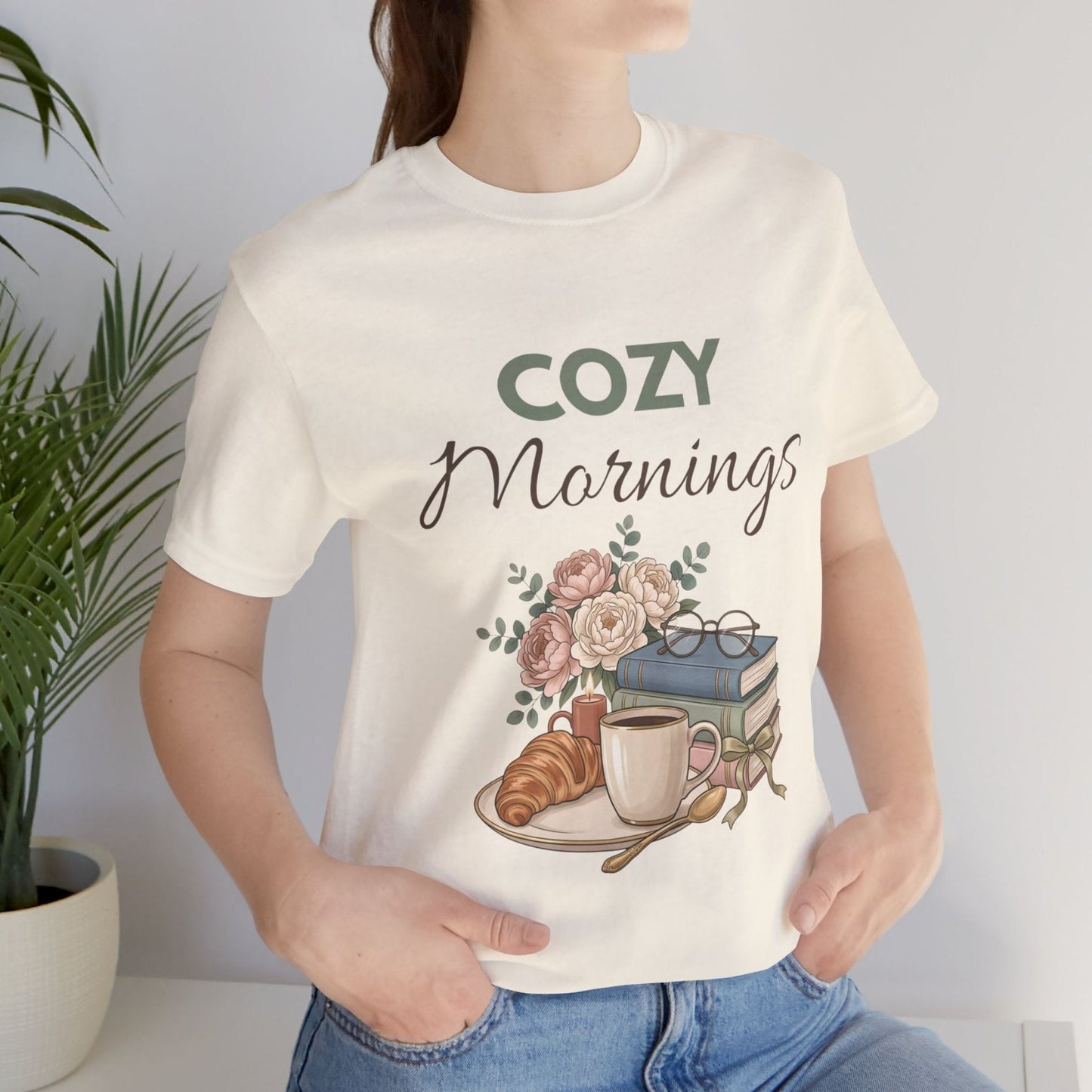 Person wearing a natural colored t-shirt with 'Cozy Mornings' design featuring a cup, croissant, and flowers.