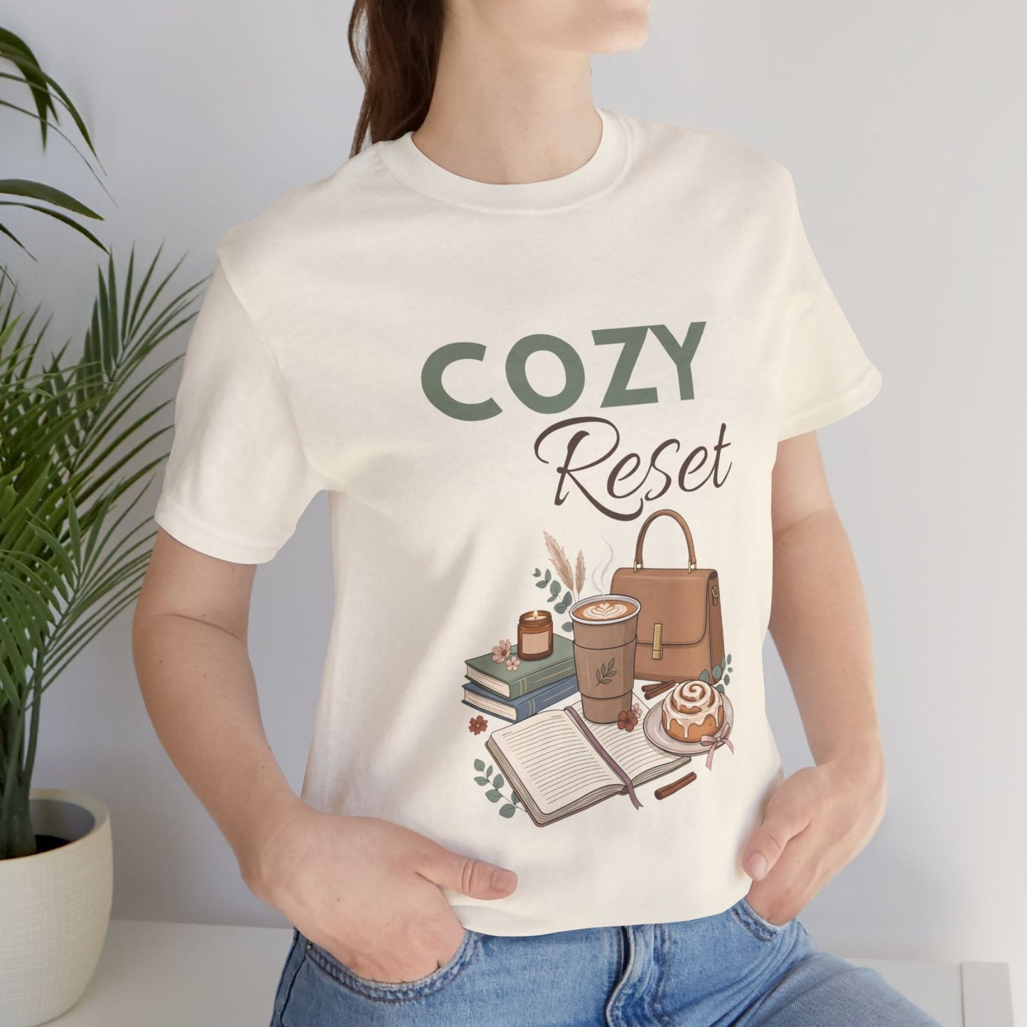 A woman is standing near a plant with a natural colored t-shirt with 'COZY Reset' on the front.