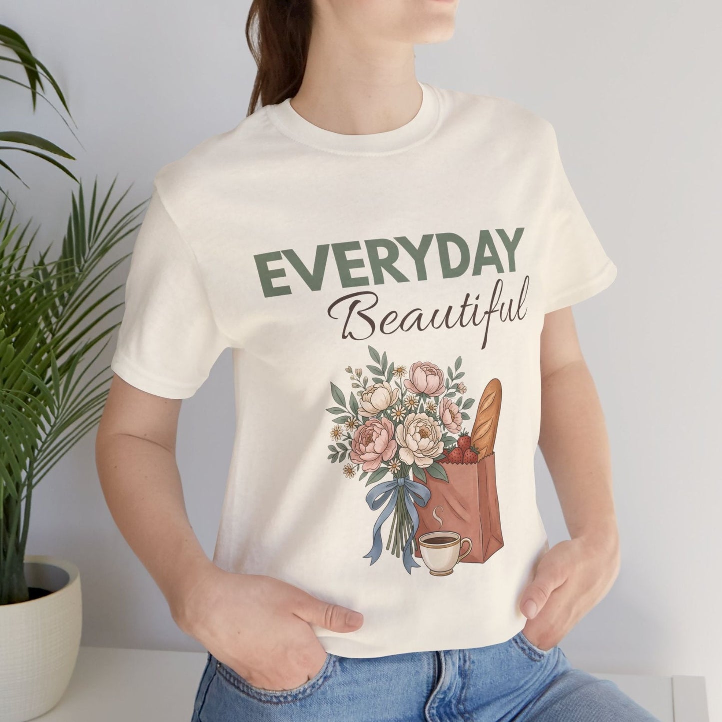 A woman standing by a plant while wearing a natural colored t-shirt with 'EVERYDAY Beautiful' on the front.