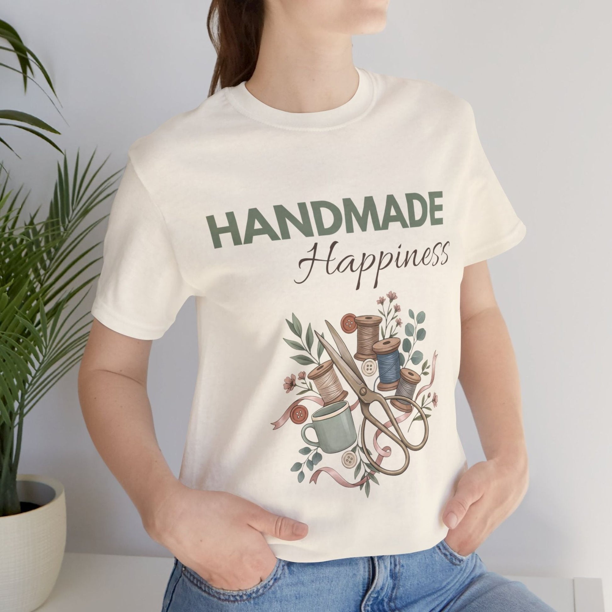 A woman standing by a plant while wearing a natural colored t-shirt with 'HANDMADE Happiness' on the front.