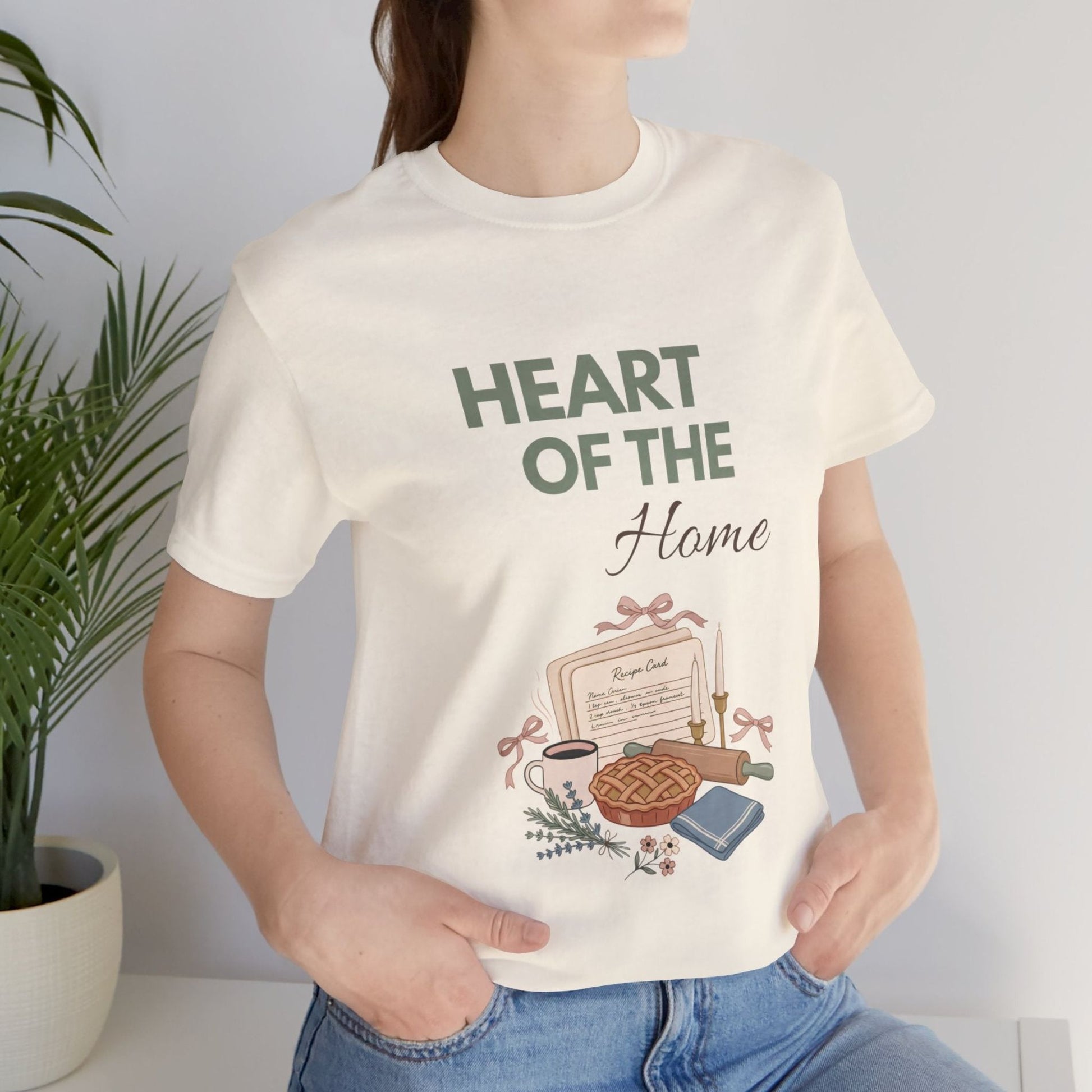 A woman is wearing a natural colored t-shirt with 'HEART OF THE Home' on the front.