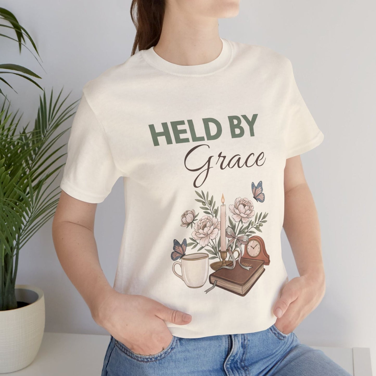 Person wearing a natural colored t-shirt with 'HELD BY Grace' design, standing against a plain background.