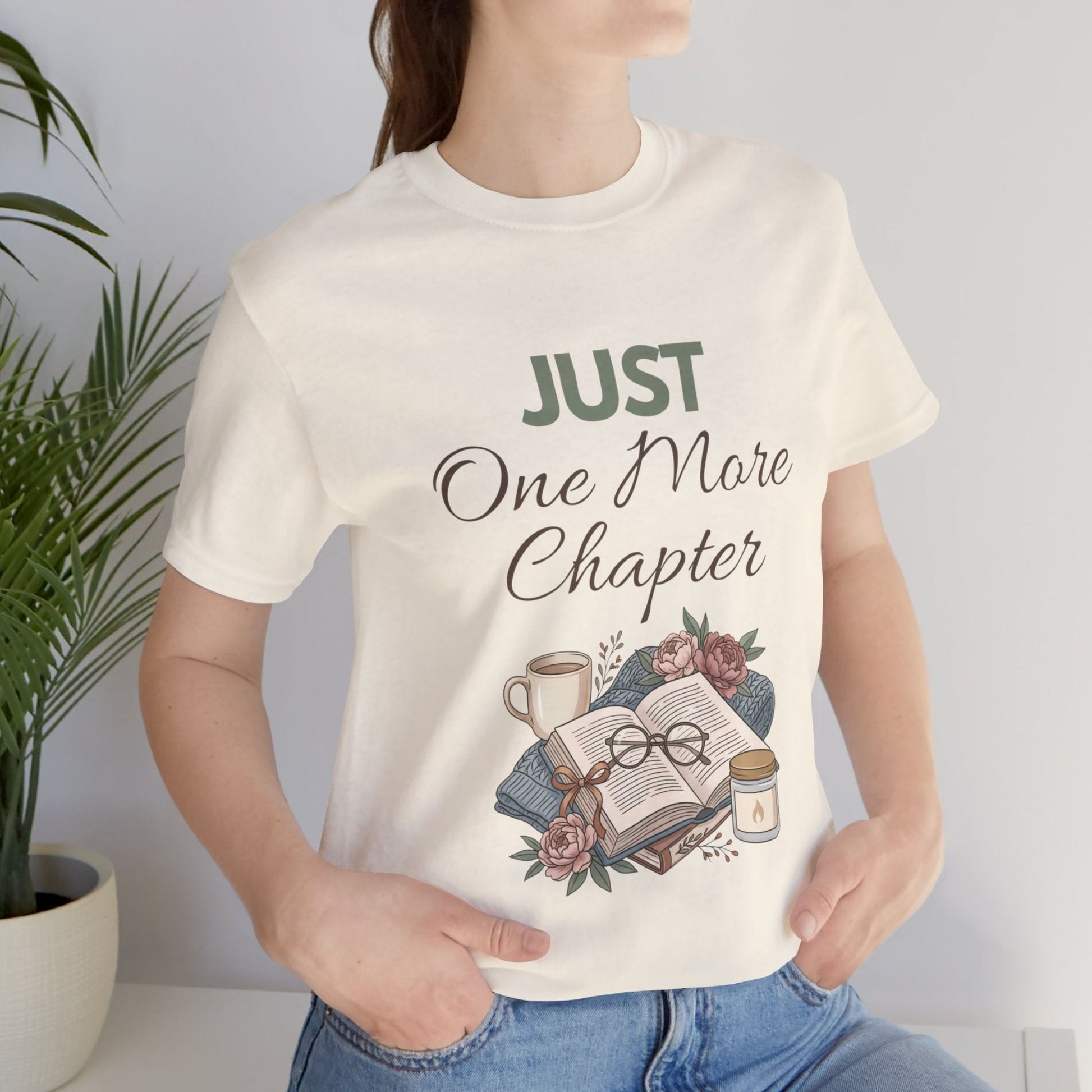 Person wearing a natural colored t-shirt with 'Just One More Chapter' design, holding a book and mug.