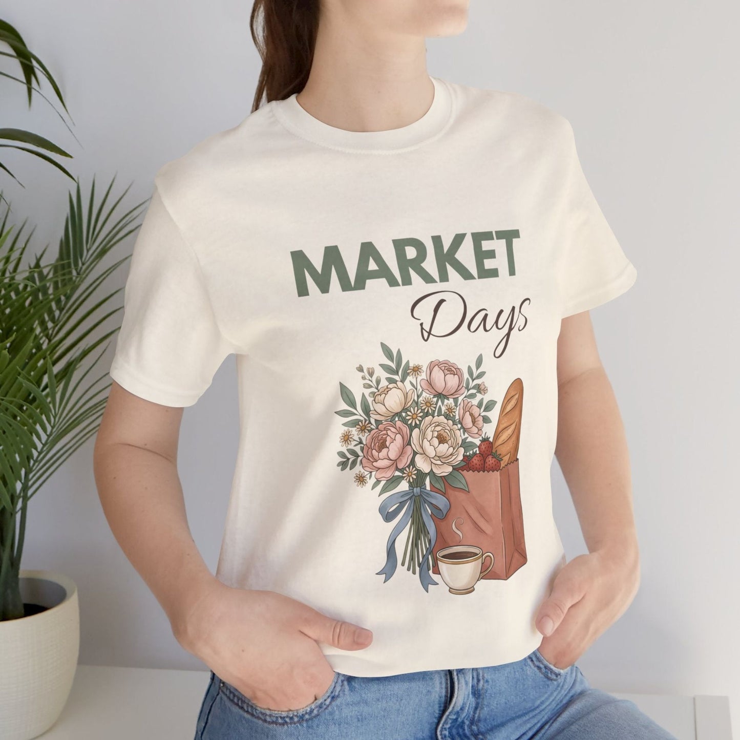 A woman wearing a natural colored t-shirt with 'Market Days' on the front standing by a plant.