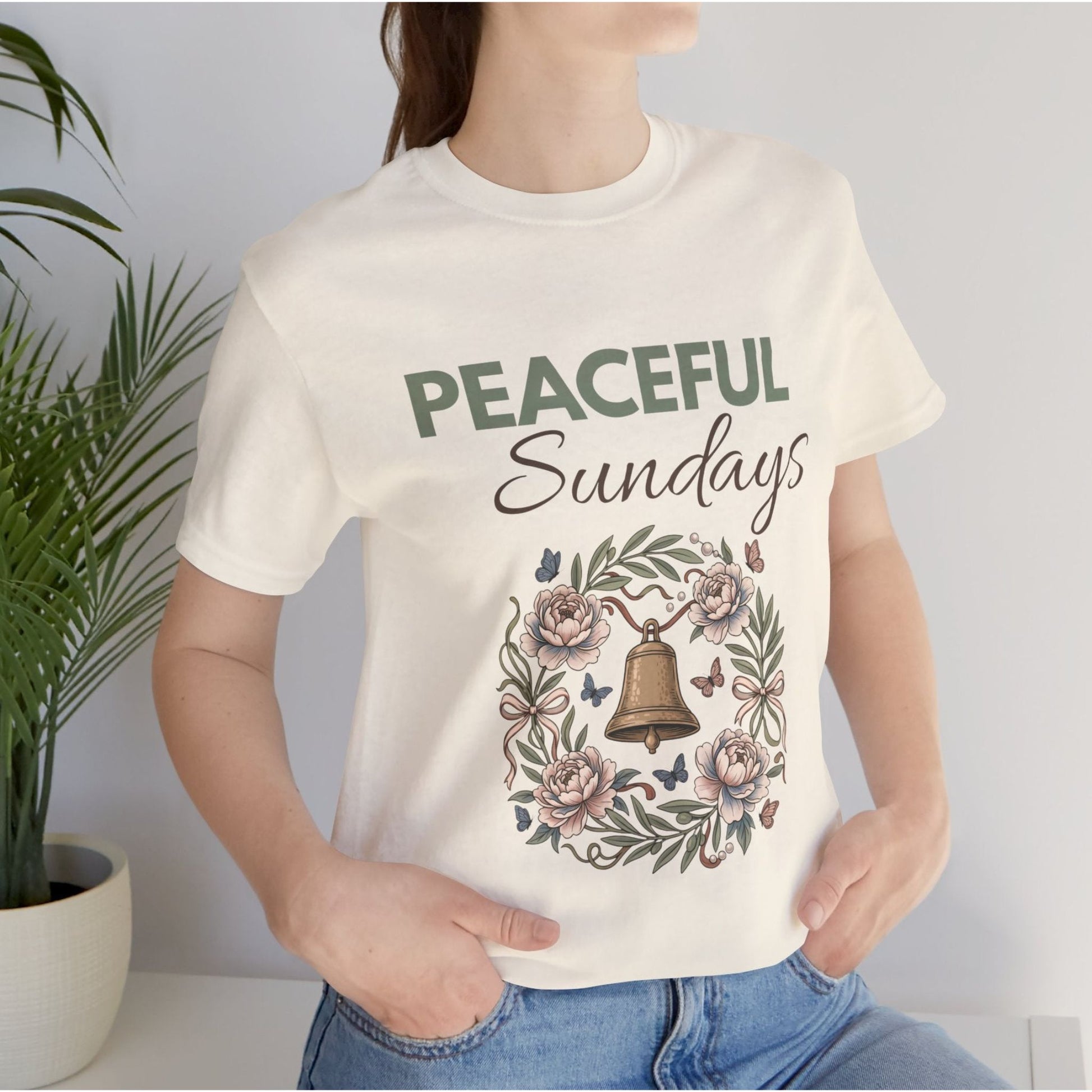 A person wearing a natural colored t-shirt with 'Peaceful Sundays' design, standing next to a plant.