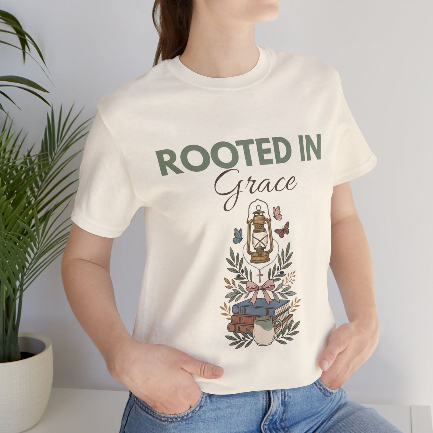 Person wearing a natural colored t-shirt with 'Rooted in Grace' design, standing next to a plant.
