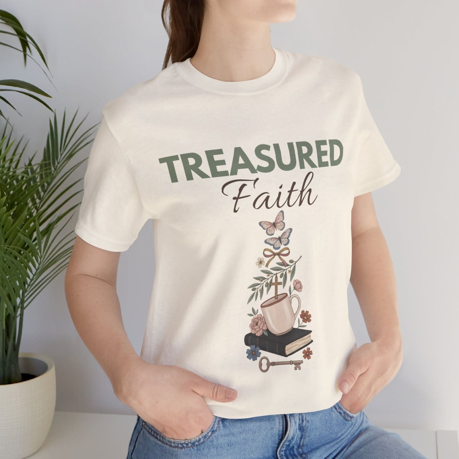 Person wearing a natural colored t-shirt with 'TREASURED Faith' design, standing next to a plant.