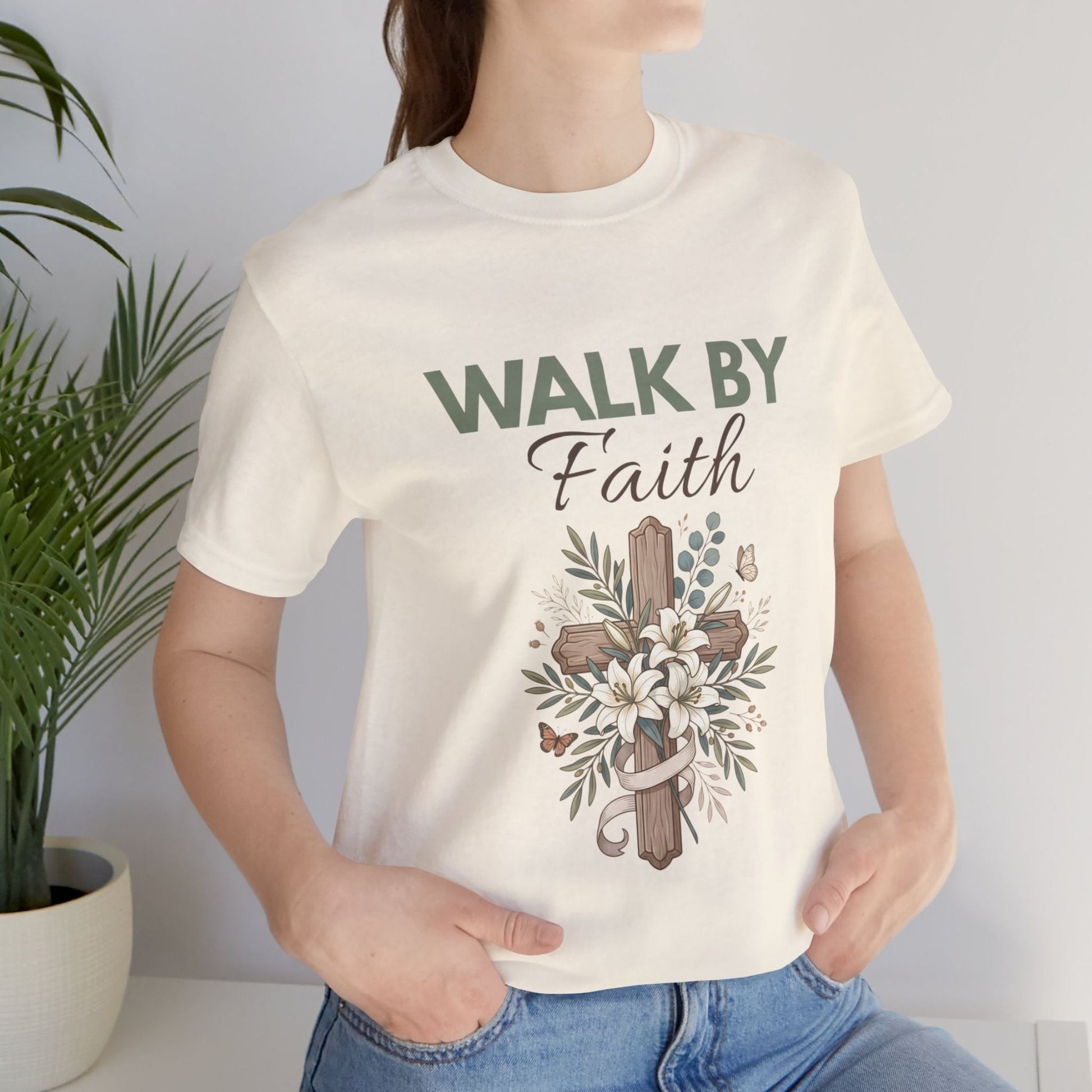 Person wearing a natural colored t-shirt with 'Walk by Faith' design, standing next to a plant.