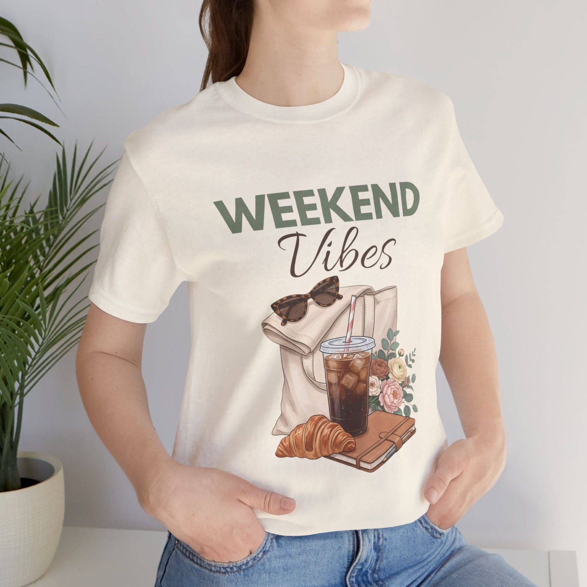 Person wearing a natural colored t-shirt with 'Weekend Vibes' design featuring a coffee cup, croissant, and sunglasses.