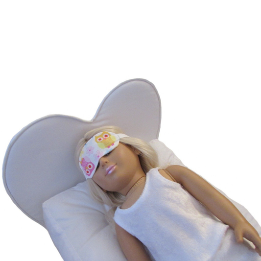 An 18-inch doll wearing a floral eye mask while lying on a white heart doll bed.