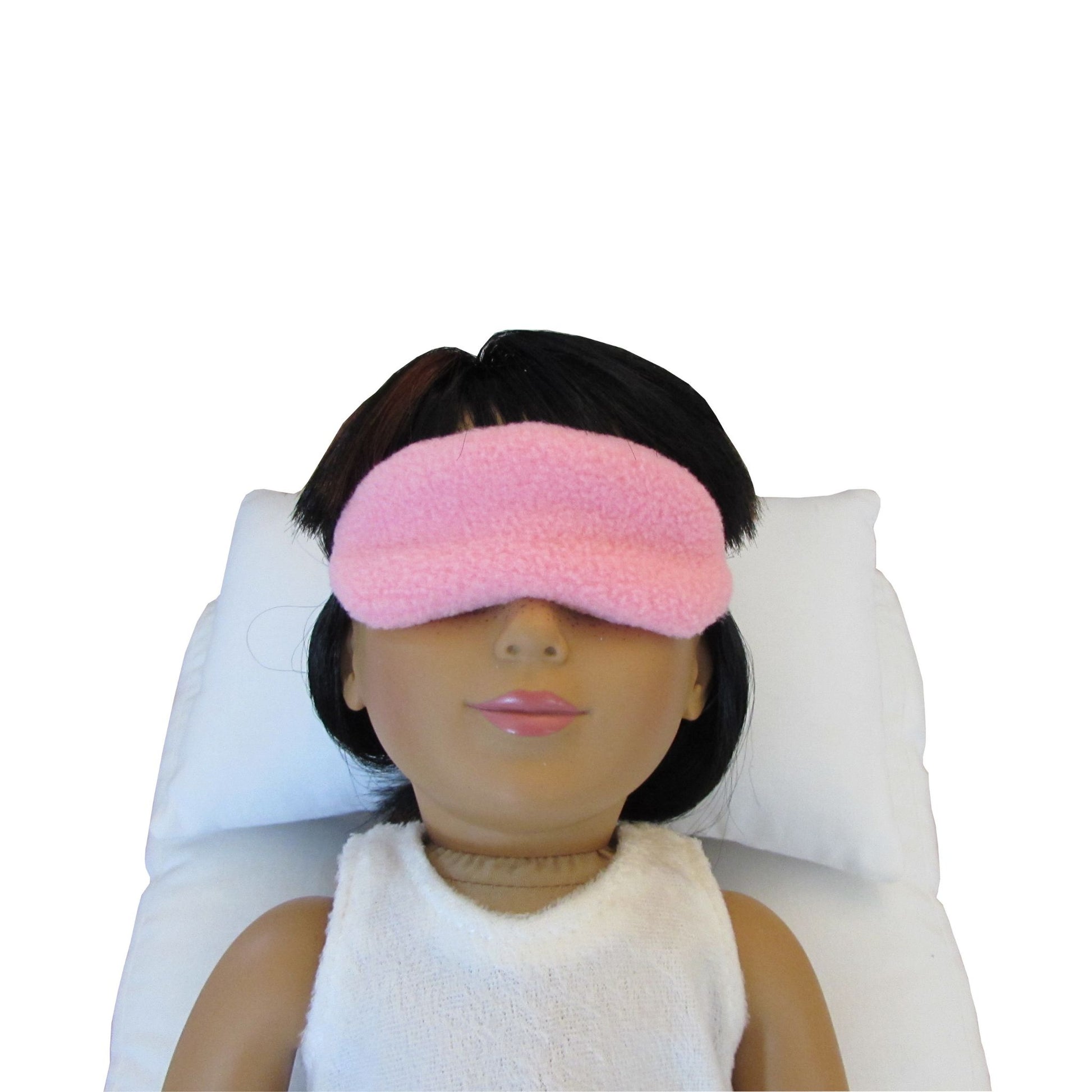 An 18-inch doll is wearing a pink anti-pill fleece doll sleep mask while lying on a white doll pillow and a white doll mattress.